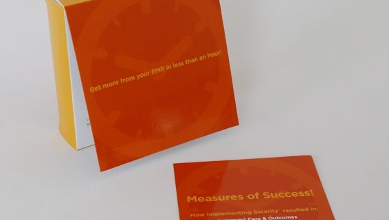 A standing bright orange clock box with a yellow back next to an orange informational paper that says measures of success