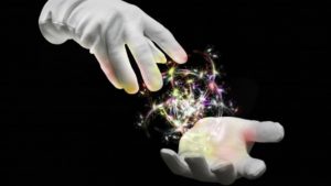 Two white gloved magician hands with a ball of multi colored sparks in between them against a dark black background