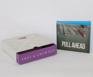 Light grey and purple slipcase box next to a light blue flap box with a picture of two boats and rowers that says Pull Ahead