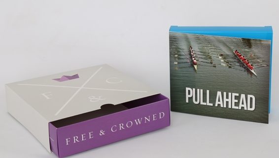 Light grey and purple slipcase box next to a light blue flap box with a picture of two boats and rowers that says Pull Ahead