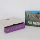 Light grey and purple slipcase box next to a light blue flap box with a picture of two boats and rowers that says Pull Ahead