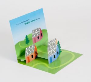 Pop up mailer opened to show cartoon style drawings of green hills with pop up houses and a bright blue sky