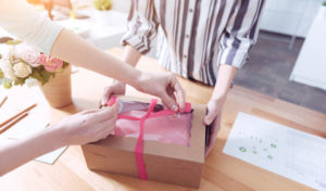 Two people assembling product packaging, one holding a brown cardboard box, the other is tying a pink ribbon around it.