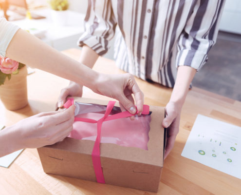 Two people assembling product packaging, one holding a brown cardboard box, the other is tying a pink ribbon around it.