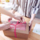 Two people assembling product packaging, one holding a brown cardboard box, the other is tying a pink ribbon around it.